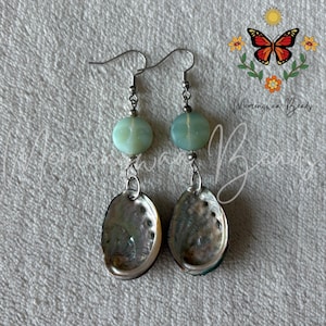 May include: A pair of dangle earrings featuring a round, pale green bead with a carved moon face design, suspended above an iridescent, oval-shaped abalone shell pendant. The earrings are attached to silver-toned metal hooks for pierced ears.