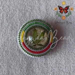 May include: A circular beaded accessory featuring a central clear resin dome containing a green cannabis leaf. The border consists of concentric rings of small green, yellow, and red beads, with a metallic silver finish on the outer edge.