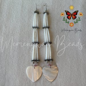 Peut inclure: Boucles d'oreilles pendantes composées de perles blanches et de pendentifs en forme de cœur en nacre. Les boucles d'oreilles ont des crochets argentés et sont ornées de perles grises et roses. De Merengwaa Beads.