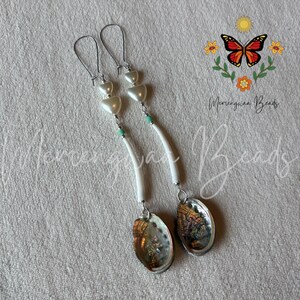 May include: A pair of long drop earrings featuring silver-tone ear wires, two white heart-shaped beads, a small turquoise bead, a curved white tube bead, and a dangling iridescent abalone shell pendant with a polished, multi-colored finish.