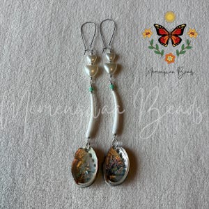 May include: A pair of long dangle earrings featuring silver-tone ear wires, two stacked white heart-shaped beads, a small turquoise accent bead, a curved white tube bead, and a round iridescent abalone shell pendant at the bottom of each.