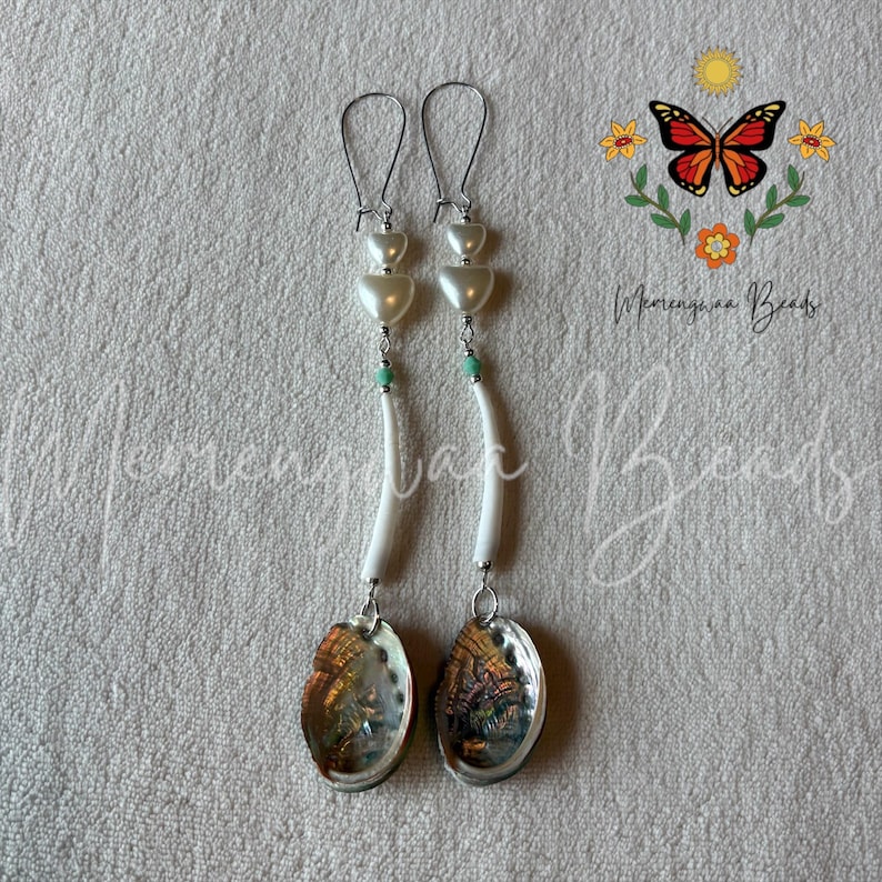 May include: A pair of long dangle earrings featuring silver-tone ear wires, two stacked white heart-shaped beads, a small turquoise accent bead, a curved white tube bead, and a round iridescent abalone shell pendant at the bottom of each.