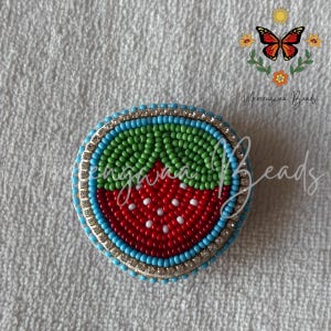 May include: A circular beaded patch featuring a strawberry design with red, green, and white seed beads. The center displays a red strawberry with white accents and a green top, surrounded by a border of light blue beads and a ring of clear rhinestones.