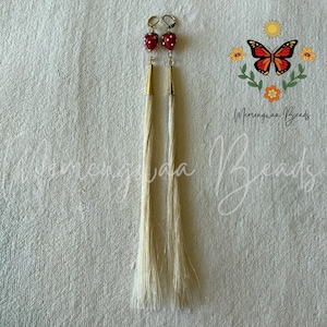 Peut inclure: Une paire de longues boucles d'oreilles pendantes avec des perles rouges en forme de fraise à pois blancs, des embouts en métal doré et de longues franges de cheveux raides blanc cassé. Le design inclut un logo papillon et du texte.