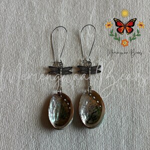 May include: A pair of dangle earrings featuring silver-toned dragonfly charms suspended from long wire hooks. Each earring is finished with an oval-shaped, iridescent abalone shell pendant that displays swirling green, blue, and purple tones.
