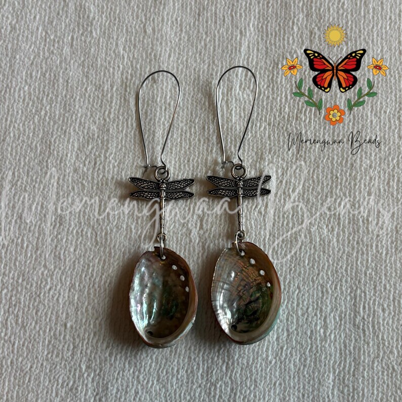 May include: A pair of dangle earrings featuring silver-toned dragonfly charms suspended from long wire hooks. Each earring is finished with an oval-shaped, iridescent abalone shell pendant that displays swirling green, blue, and purple tones.