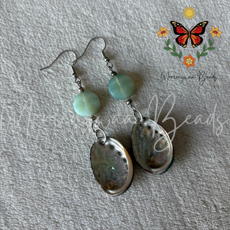 May include: Dangle earrings featuring a round, pale green bead with a carved face design suspended above an oval-shaped, iridescent abalone shell pendant. The components are connected by silver-toned metal findings and hooks.