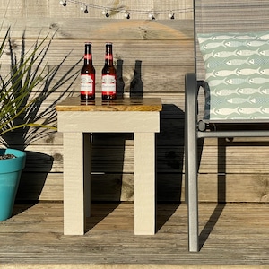Könnte beinhalten: Ein kleiner, weißer Holz-Beistelltisch mit einer natürlichen Holzplatte. Zwei Flaschen Bier stehen auf dem Tisch. Der Tisch steht auf einem Holzdeck mit einem Stuhl und einer Pflanze in einem blauen Topf.