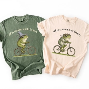 May include: Two t-shirts, one in green and one in beige, each with a playful design of a frog on a bicycle. The green shirt shows a frog in a wizard hat with the text "off to commit uncle foolery." The beige shirt has a frog with a floral crown, and the text "off to commit aunt foolery."
