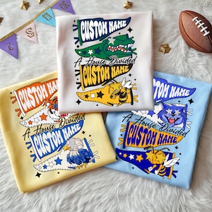 Könnte beinhalten: Vier gefaltete T-Shirts mit individuellen Designs. Die Shirts sind weiß, gelb und hellblau. Jedes Shirt hat ein Wimpeldesign mit dem Text "CUSTOM NAME" und "A House Divided". Die Designs umfassen einen Alligator, eine Biene, einen Tiger, einen Adler und eine Katze.