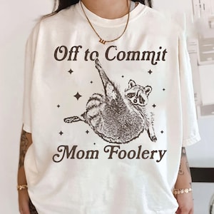 May include: Off-white t-shirt featuring the phrase "Off to Commit Mom Foolery" and a raccoon illustration. The raccoon is drawn in brown ink. The shirt is a casual wear item.