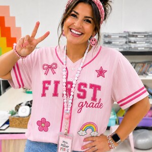 May include: A light pink baseball-style jersey with white pinstripes, featuring the text "FIRST grade" in dark pink, adorned with embroidered patches of a bow, star, flower, and rainbow, paired with blue jeans and a beaded lanyard with an ID badge.