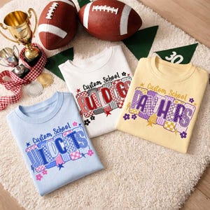 以下が含まれることがあります： ライトブルー、ホワイト、ペールイエローのクルーネックTシャツ3枚。各シャツには「Custom School」の文字とチーム名（Wildcats、Bulldogs、Panthers）がプリントされています。文字にはカラフルなチェック柄や格子柄が施されています。