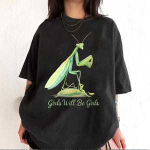 Może przedstawiać: Czarny t-shirt z akwarelową ilustracją zielonej modliszki. Modliszka stoi na ż&oacute;łwiu, z napisem "Girls Will Be Girls" w zielonej czcionce poniżej.