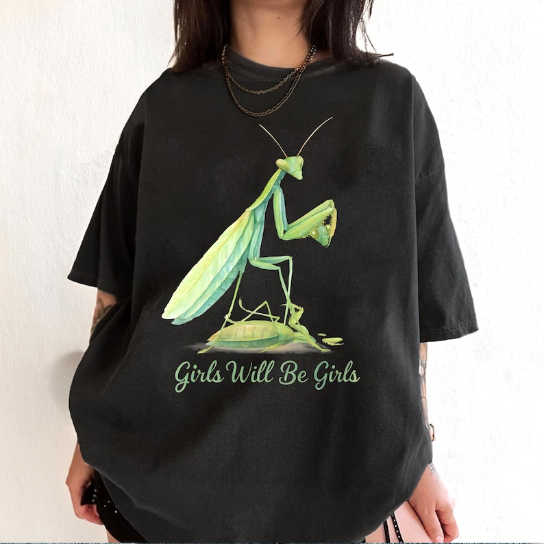 Może przedstawiać: Czarny t-shirt z akwarelową ilustracją zielonej modliszki. Modliszka stoi na ż&oacute;łwiu, z napisem "Girls Will Be Girls" w zielonej czcionce poniżej.