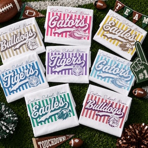 Peut inclure: Une collection de sept t-shirts blancs avec des rayures verticales de différentes couleurs, chacun imprimé avec un nom d'école personnalisé, un nom de mascotte comme Gators, Eagles, Tigers ou Bulldogs, et une illustration associée.