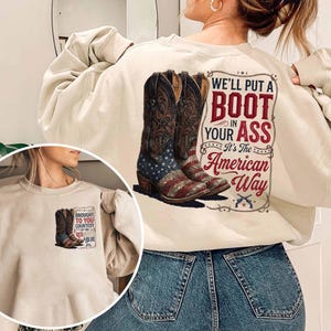 Puede incluir: Sudadera color crema con un gráfico de botas de vaquero con diseño de bandera estadounidense y el texto "WE'LL PUT A BOOT IN YOUR ASS It's The American Way." Un gráfico más pequeño de una bota está en el frente.