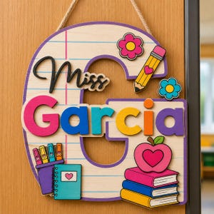 May include: A wooden door sign shaped like the letter G with a lined paper pattern. It features the black script text Miss Garcia, a yellow pencil, a pink flower, a blue flower, a purple pencil holder, a teal notebook, and a stack of books with an apple.