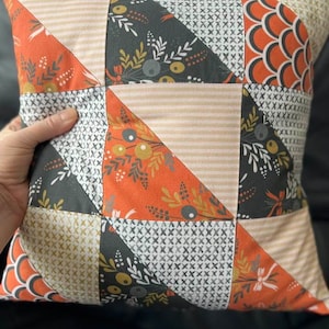 May include: A patchwork pillow with a geometric design featuring orange, black, white, and beige fabric. The pillow is made of different fabrics with various patterns, including floral, geometric, and striped.