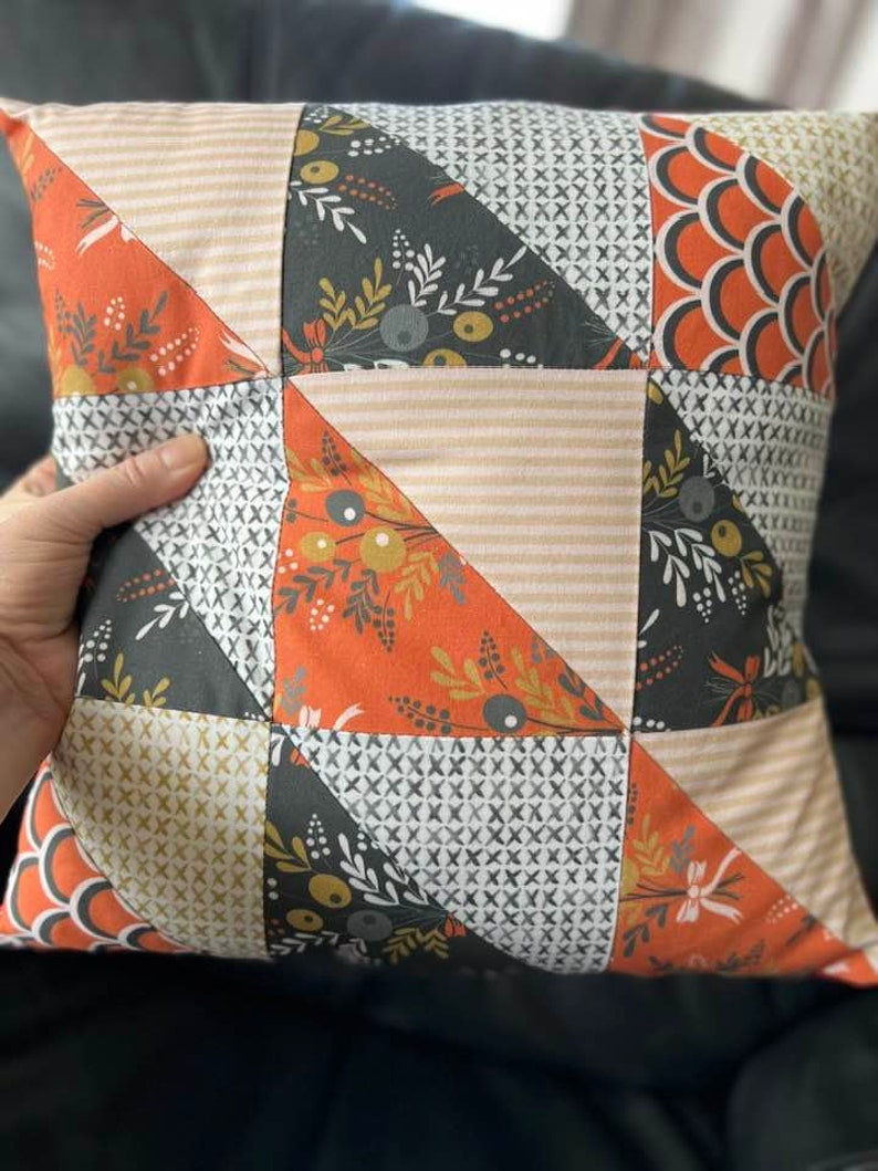 May include: A patchwork pillow with a geometric design featuring orange, black, white, and beige fabric. The pillow is made of different fabrics with various patterns, including floral, geometric, and striped.
