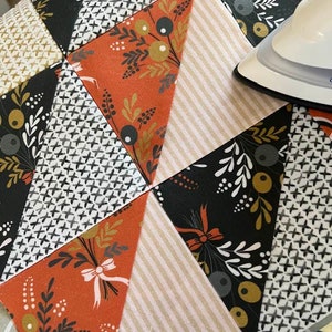 May include: A close-up of a quilt being ironed. The quilt is made of various fabrics with different patterns, including floral, geometric, and striped designs. The colors are predominantly black, white, orange, and brown.
