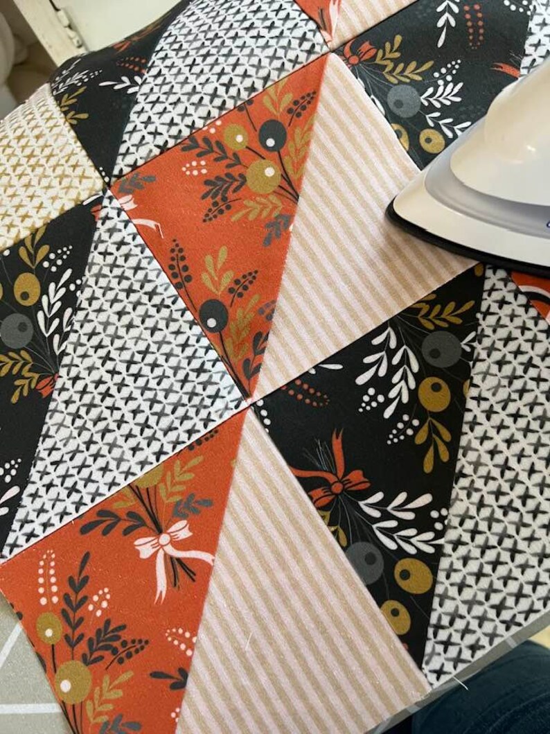 May include: A close-up of a quilt being ironed. The quilt is made of various fabrics with different patterns, including floral, geometric, and striped designs. The colors are predominantly black, white, orange, and brown.