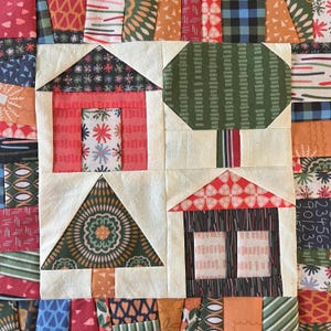May include: A quilt block with a patchwork design featuring a green tree, a red house, a red house with a white roof, and a brown tree. The quilt block is made of various fabrics with different patterns and colors.