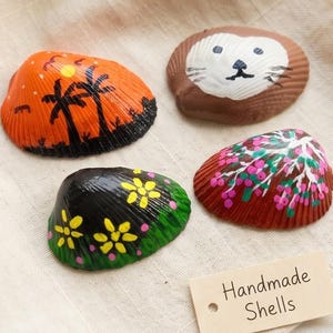 May include: Four hand-painted seashells with unique designs. One shell features a sunset scene with palm trees, another a lion face, a third with yellow flowers, and the last with pink flowers. The tag reads "Handmade Shells."