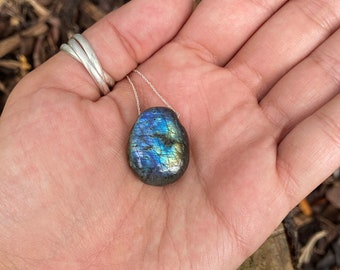 May include: A teardrop-shaped labradorite pendant necklace featuring a polished stone with iridescent blue and gold flashes. The pendant is suspended from a delicate silver-colored chain, showcasing the natural mineral's reflective surface.