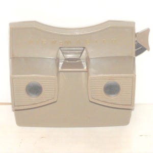 May include: Tan View-Master viewer with the word "VIEW MASTER" printed across the top. The viewer has two circular eye holes and a lever on the side. The design is a classic, retro style.