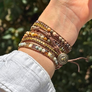 Könnte beinhalten: Wickelarmband mit mehreren Strängen aus Perlen in Erdtönen, darunter Braun, Gold und Grün. Das Armband wird mit einem Knopfverschluss und einem Lederband befestigt. Das Design beinhaltet verschiedene Texturen und Größen von Perlen.
