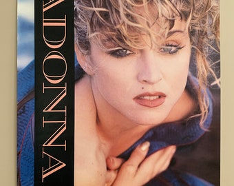 May include: A vintage poster featuring a close-up portrait of Madonna. The poster has a black background with the word "MADONNA" in white lettering. The poster lists the songs "Borderline", "Holiday", "Burning Up", "Lucky Star", "Like a Virgin", "Material Girl", and "Angel".