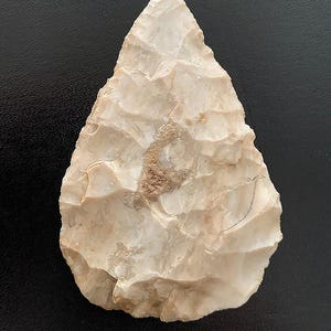 May include: A hand-knapped, off-white stone arrowhead with a triangular shape. The arrowhead has a textured surface with visible chipping and a small area of brown discoloration. The artifact is likely an ancient tool.