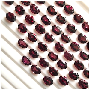 Puede incluir: Una colecci&oacute;n de 40 piedras preciosas ovaladas de color rojo oscuro. Las piedras preciosas est&aacute;n dispuestas en filas sobre una superficie blanca.