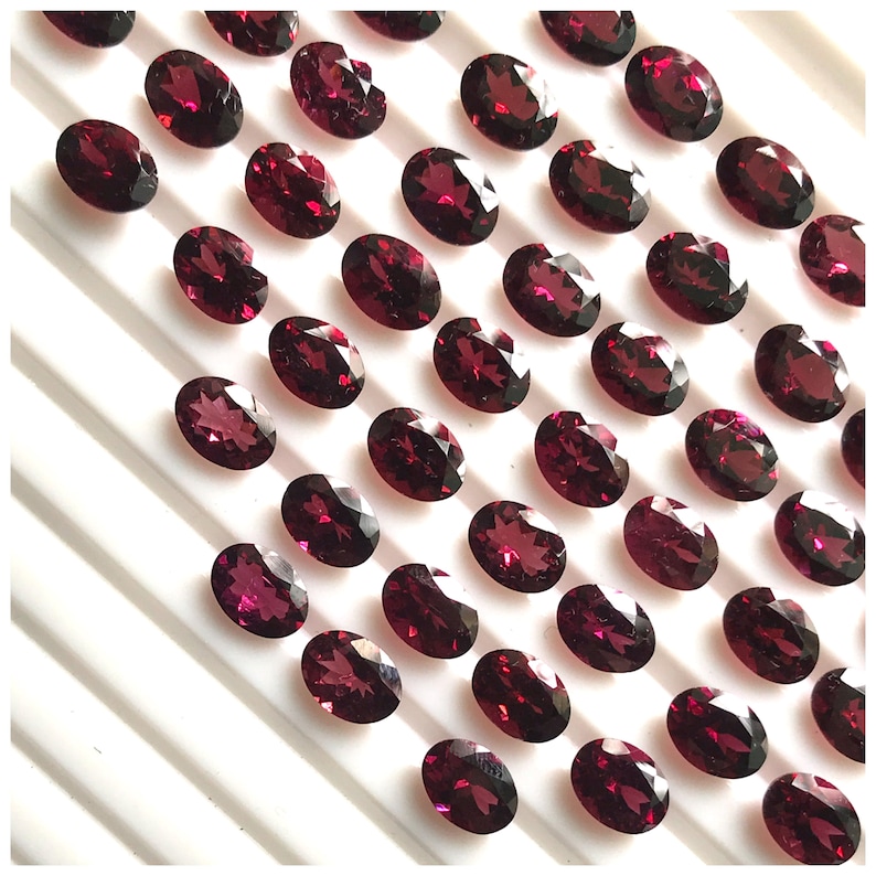 Puede incluir: Una colecci&oacute;n de 40 piedras preciosas ovaladas de color rojo oscuro. Las piedras preciosas est&aacute;n dispuestas en filas sobre una superficie blanca.