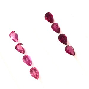 May include: Eight pear-shaped pink gemstones arranged in two rows on a white surface.