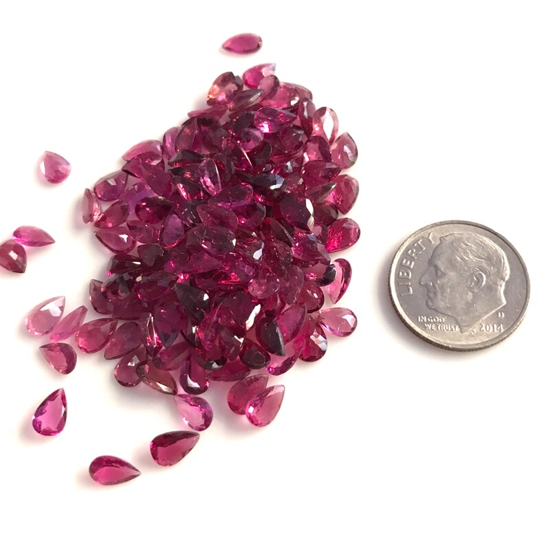 May include: A large pile of pink pear-shaped gemstones, with a few scattered around, next to a UK ten pence coin for scale.