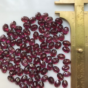 Puede incluir: Un mont&oacute;n de aproximadamente 60 peque&ntilde;as piedras preciosas talladas de forma ovalada. Las piedras preciosas son de color p&uacute;rpura rojizo oscuro. Una regla est&aacute; en la imagen para la escala. La regla est&aacute; marcada en pulgadas y cent&iacute;metros.