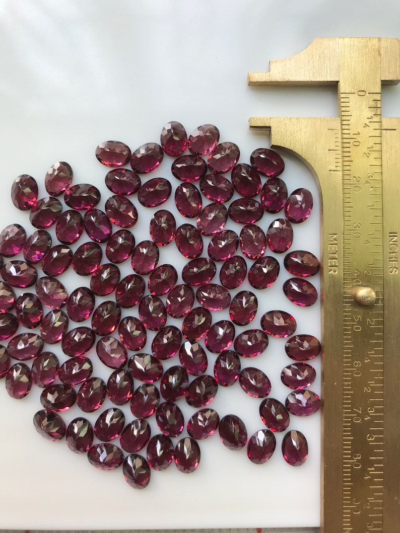 Puede incluir: Un mont&oacute;n de aproximadamente 60 peque&ntilde;as piedras preciosas talladas de forma ovalada. Las piedras preciosas son de color p&uacute;rpura rojizo oscuro. Una regla est&aacute; en la imagen para la escala. La regla est&aacute; marcada en pulgadas y cent&iacute;metros.