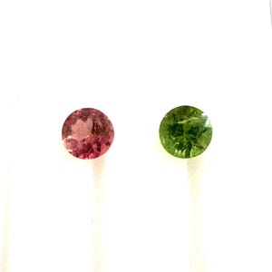May include: Two round gemstones, one pink and one green, are shown against a white backdrop. The pink gemstone has a reddish-pink hue, while the green gemstone has a vibrant, verdant colour. Both stones appear to be faceted.