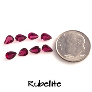 May include: Ten pear-shaped rubellite gemstones, each approximately the size of a ten pence coin, are arranged in two rows on a white surface. The gemstones are a deep pink colour and have a slight sparkle. A UK ten pence coin is included for size comparison.
