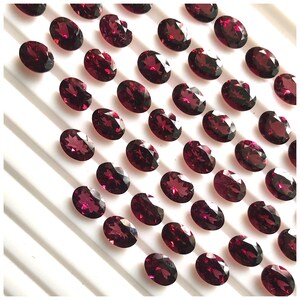 Puede incluir: Una colecci&oacute;n de 40 piedras preciosas ovaladas de color rojo oscuro. Las piedras preciosas est&aacute;n dispuestas en filas sobre una superficie blanca.