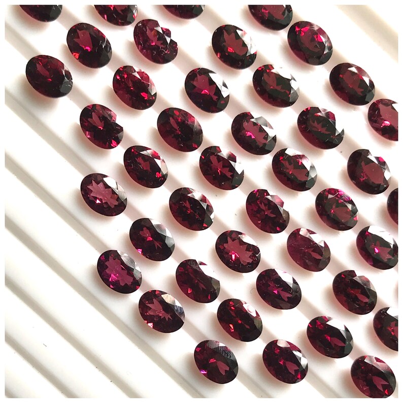 Puede incluir: Una colecci&oacute;n de 40 piedras preciosas ovaladas de color rojo oscuro. Las piedras preciosas est&aacute;n dispuestas en filas sobre una superficie blanca.