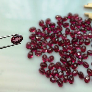Puede incluir: Primer plano de un mont&oacute;n de peque&ntilde;as piedras preciosas rojas de forma ovalada. Una piedra preciosa se sostiene con unas pinzas, mostrando sus facetas.