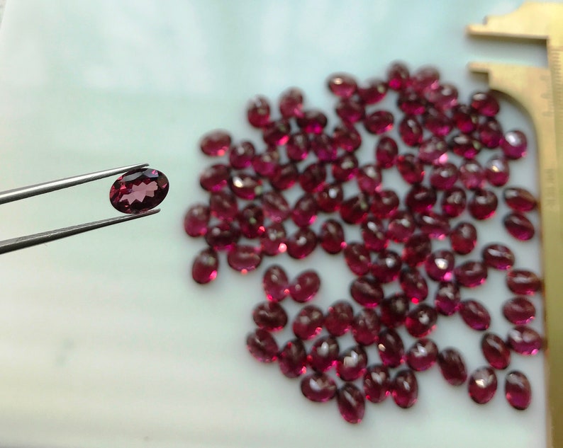 Puede incluir: Primer plano de un mont&oacute;n de peque&ntilde;as piedras preciosas rojas de forma ovalada. Una piedra preciosa se sostiene con unas pinzas, mostrando sus facetas.