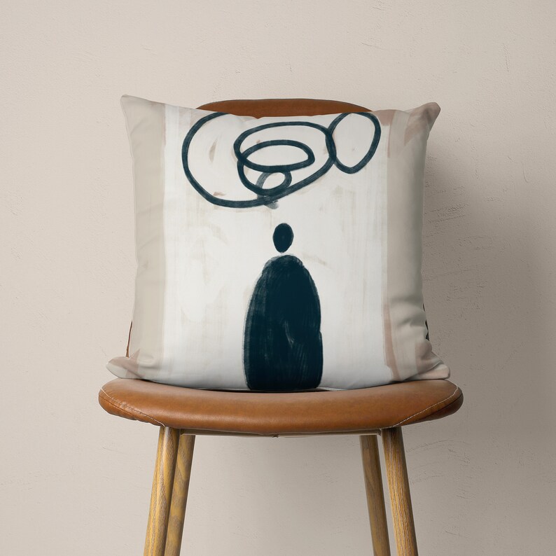 Puede incluir: Una almohada con un fondo beige y un dise&ntilde;o abstracto negro. El dise&ntilde;o presenta una forma de remolino y una figura azul oscuro.