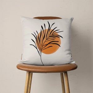May include: A white throw pillow with a black and orange abstract design featuring a stylized palm leaf and a circle.