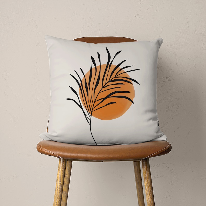 May include: A white throw pillow with a black and orange abstract design featuring a stylized palm leaf and a circle.