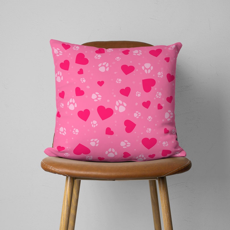 May include: A square pink throw pillow with a pattern of bright pink hearts, white paw prints, and small white dots. The pillow is placed on a brown wooden chair, creating a cozy and inviting aesthetic.