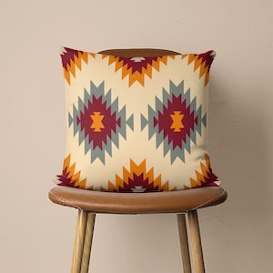 Op de afbeelding: Een decoratief kussen met een geometrisch patroon in tinten oranje, rood, grijs en beige. Het kussen ligt op een bruine stoel.
